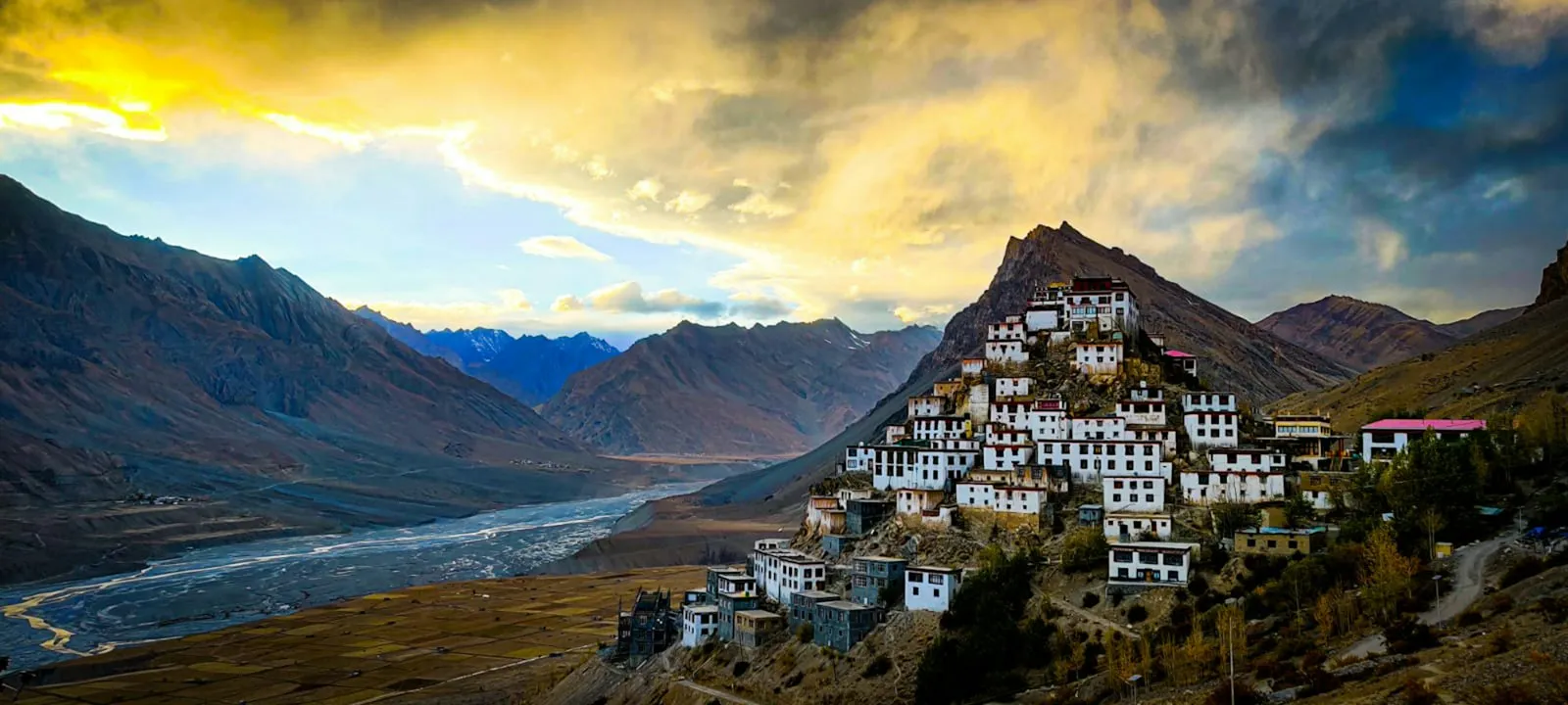 A small Spiti village of flat-roofed houses on a hillside above the grey Spiti River, ringed by high desert mountains