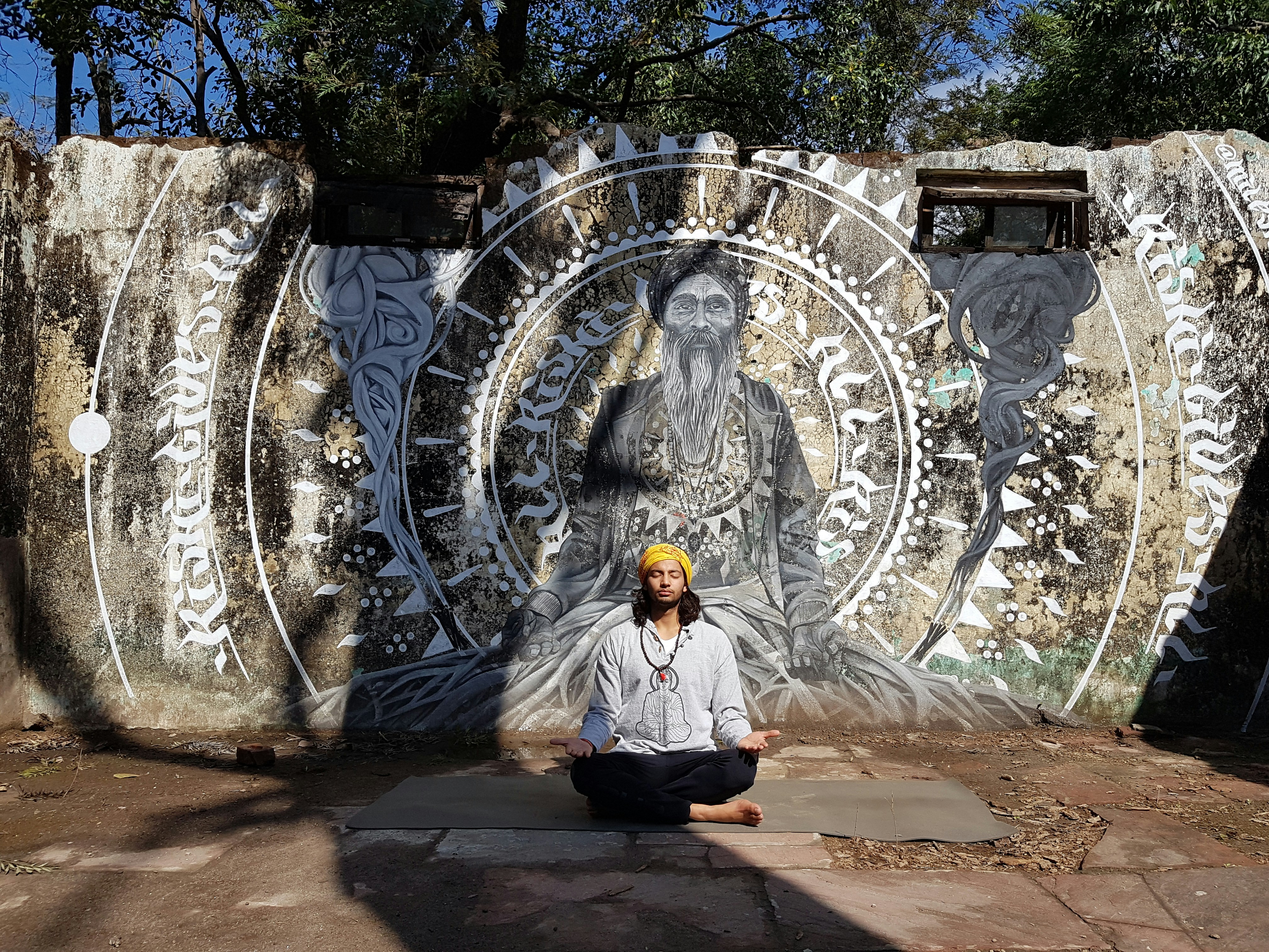 A man meditating cross-legged on a mat before a large painted mural of a sadhu in Rishikesh